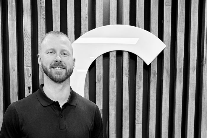 A black and white portrait of a smiling man with a beard wearing a dark polo shirt, standing in front of a slatted wooden wall with a large white arch-shaped logo behind him.